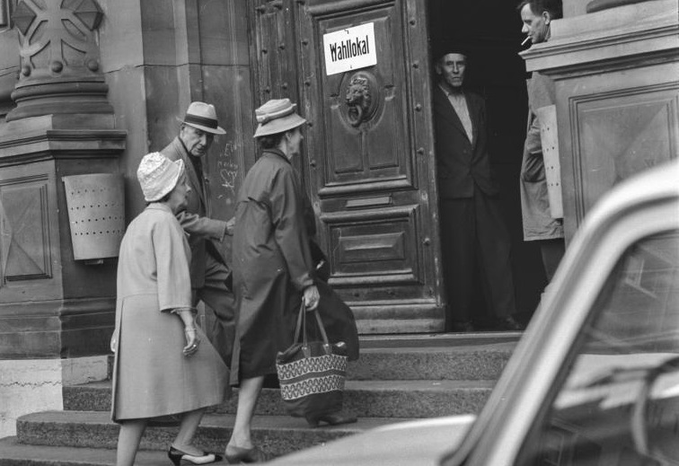 Schwarz-Weiss Foto: Zwei Frauen und ein Mann betreten ein Wahllokal.
