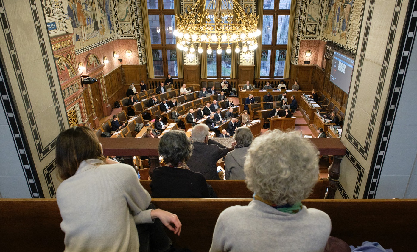 Auf der Tribüne sitzt Publikum und schaut in den Saal hinunter, wo das Parlament tagt.
