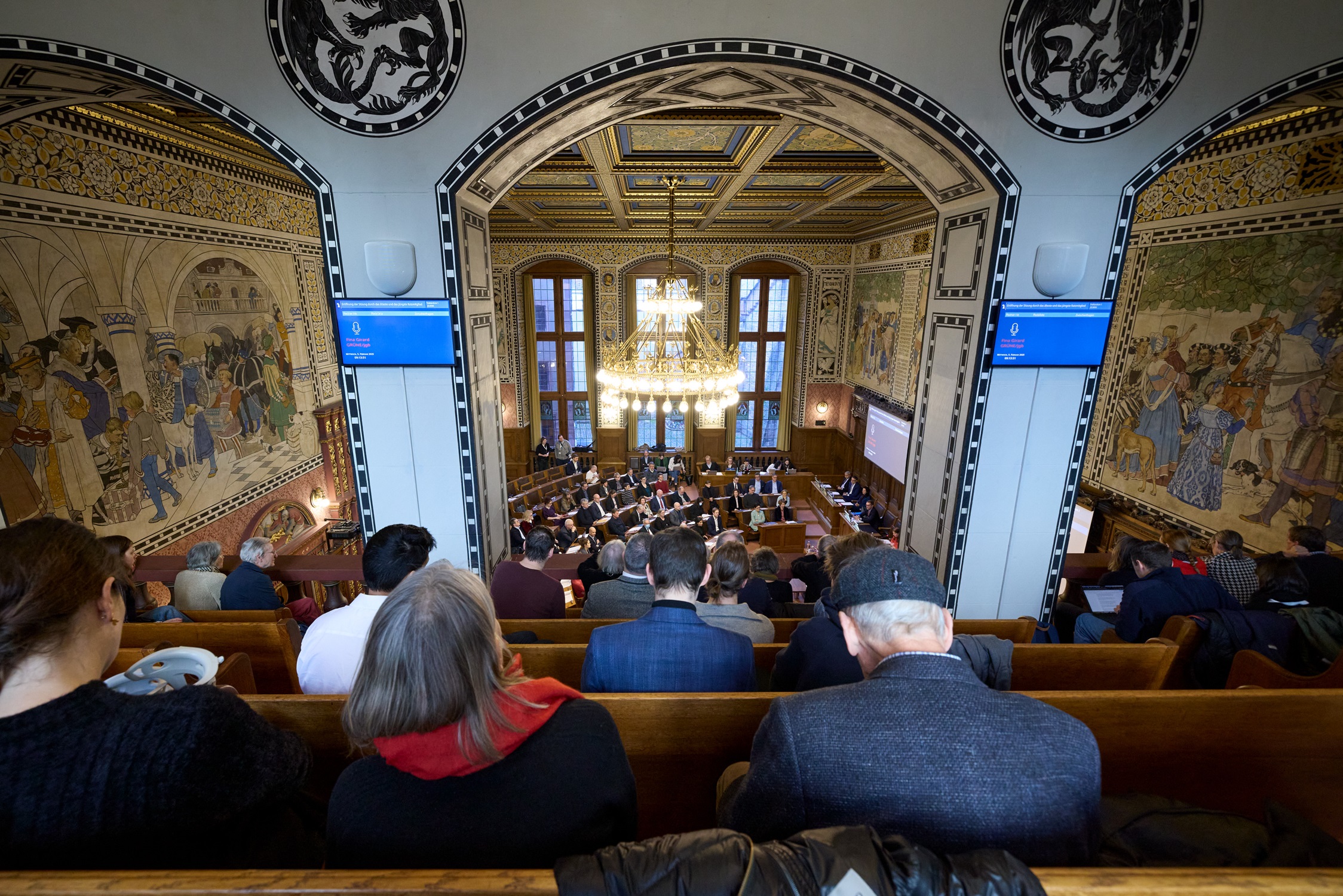 Von der gut besetzten Tribüne aus blicken Zuschauende auf den Grossen Rat in Sitzung. Er politisiert in einem wunderschönen historischen Saal im Basler Rathaus. Die Sitzungen sind öffentlich. 